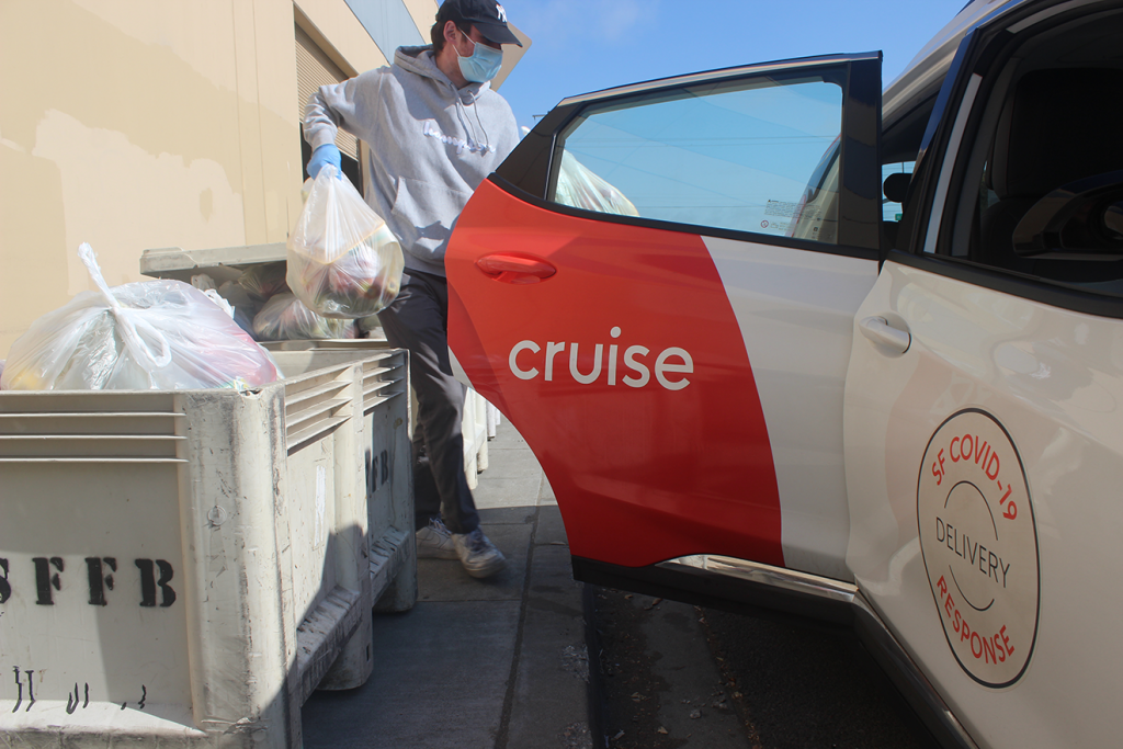 A man in a gray hoodie, mask, and baseball cap holds several plastic bags of food items which he is loading into a backseat of a car.